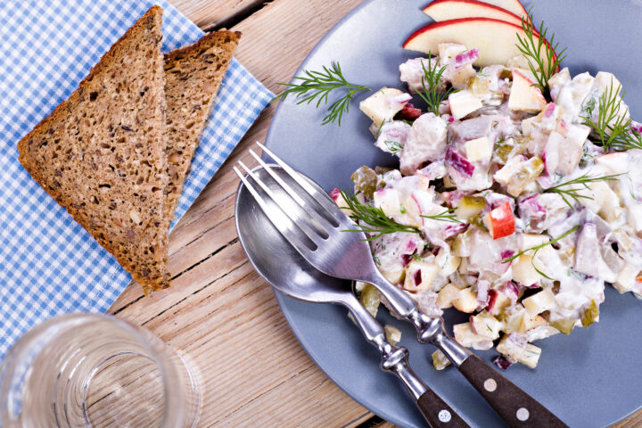 auf der rechten Abbildungsseite ist ein Teller mit einem Fischsalat, darauf sind Dillzweige, feine Apfelschnitze und das Besteck angerichtet. Daneben liegt eine karrierte Servierte und zwei Scheiben Vollkornbrot darauf. Dazu ist ein Glas Wasser zu erkennen.