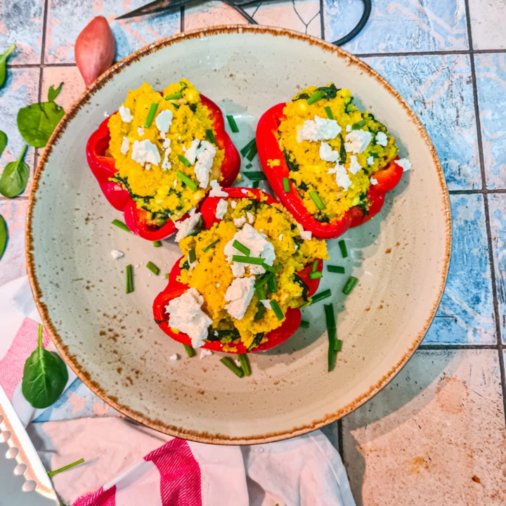 heller Teller mit drei hälften von roten Paprikas gefüllt mit Couscous, Spinat und Feta. Der Teller ist von oben fotografiert. Rechts unten neben dem Teller ist ein helles Küchenhandtuch mit roten Streifen und ein Kochmesser zu erkennen.